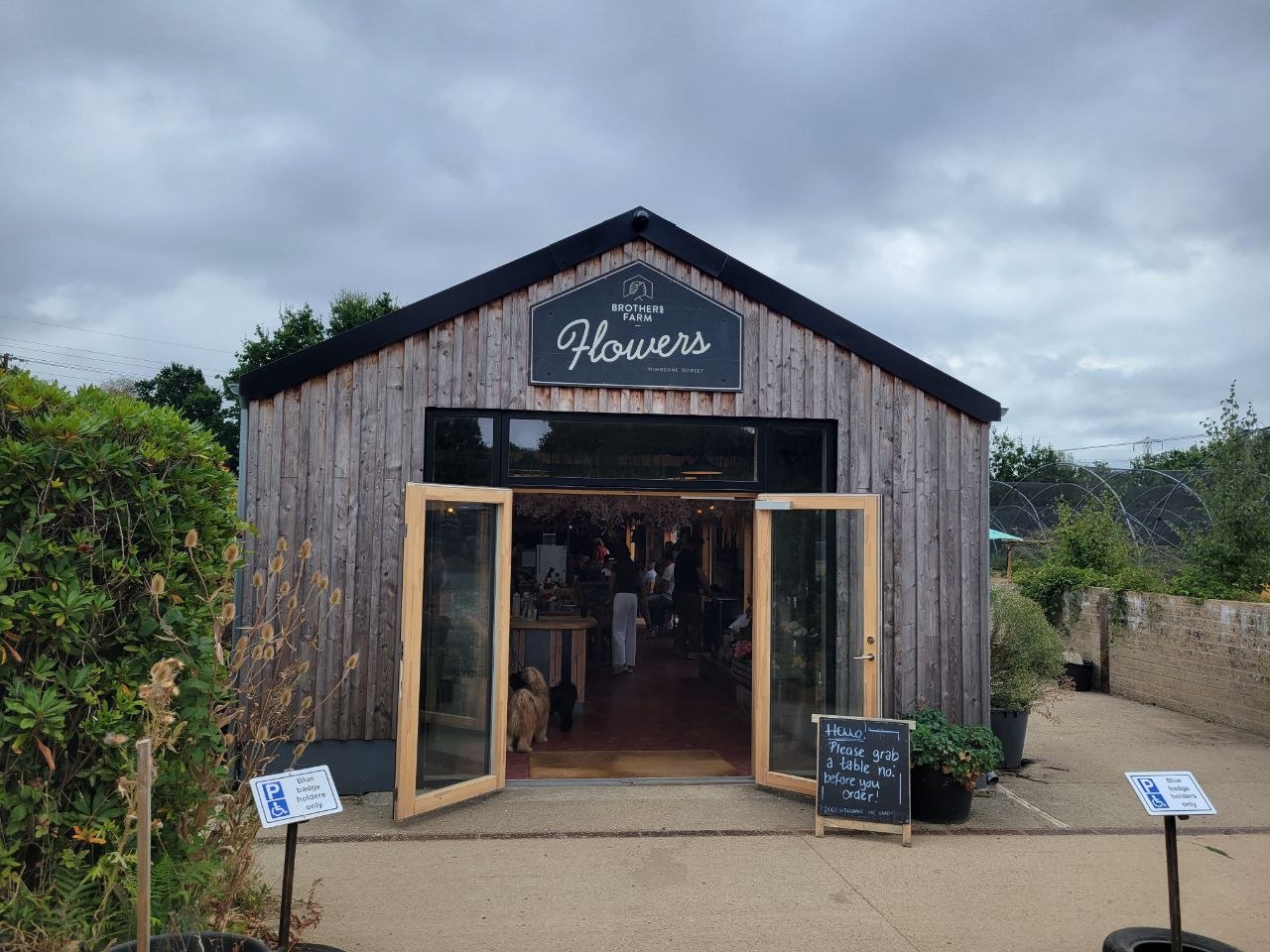 The Brothers Farm Flowers building: a bare-timber barn with a painted sign on the gable end, its glazed double doors propped open onto a level concrete forecourt, with a chalkboard and two blue disabled-parking bay signs outside