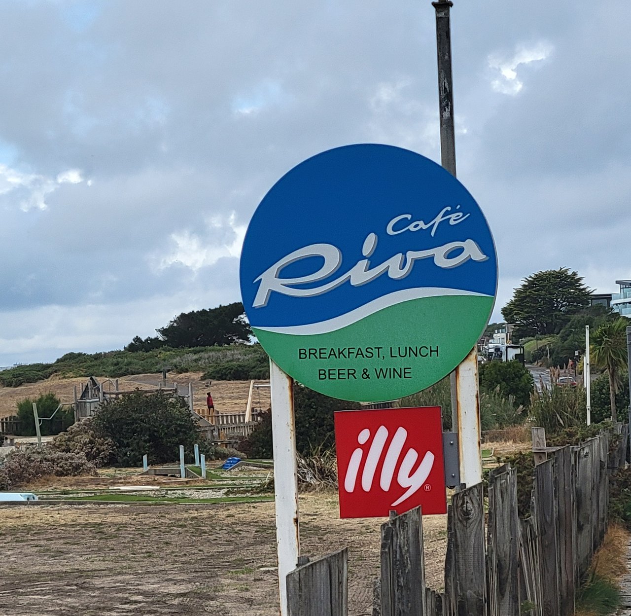 The round blue and green Café Riva sign reading Breakfast, Lunch, Beer and Wine on a post beside a wooden fence, with a red illy coffee sign below it and the clifftop green and a playground behind
