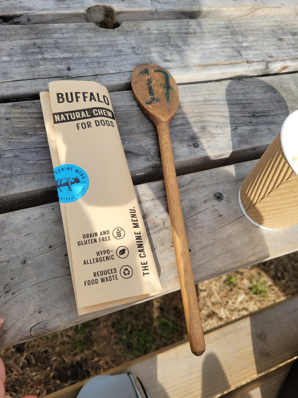 A Canine Menu buffalo natural chew for dogs in its cream paper sleeve, labelled grain and gluten free, hypoallergenic and reduces food waste, lying on a weathered wooden table next to a wooden spoon marked with a table number and a takeaway coffee cup